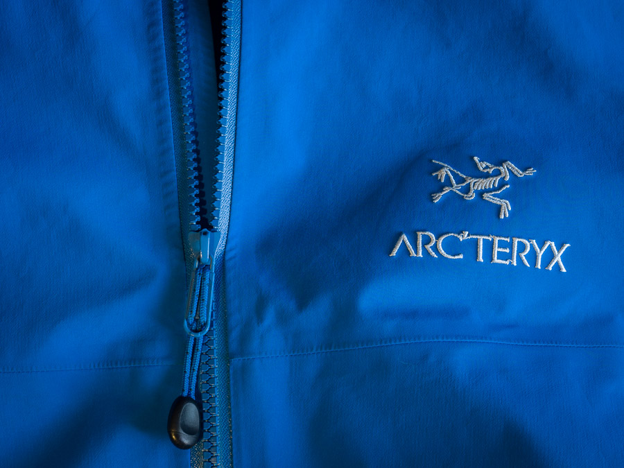 Photo of the chest of the jacket laid flat. Showing the main zip partially unzipped and the Arc'teryx logo and name embroidered in white.