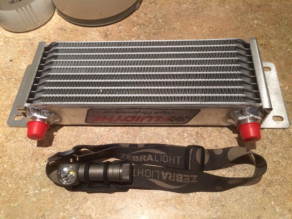 Oil cooler, with a headlamp for scale. Cooler is approximately one foot wide by four inches tall by two inches deep.